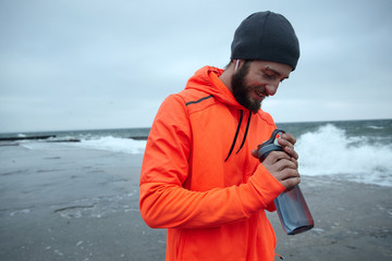 Handsome young dark haired bearded man goes in for sport every morning, going along seafront with fitness bottle in raised hands and smiling positively. Sport and healthy lifestyle concept