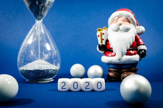 Blue Christmas Background With Santa Clause Figure And Running Hour Glass In A Conceptual Image With White Ornaments And 2020 Spelled On White Blocks