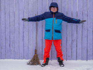 man in bright ski suit stands holding broom in his hands and is going to clean the snow in winter...