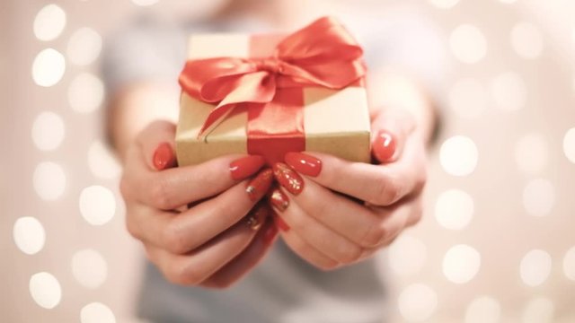 Unrecognizable young woman gives a gift box with red ribbon to camera on blurred lights.