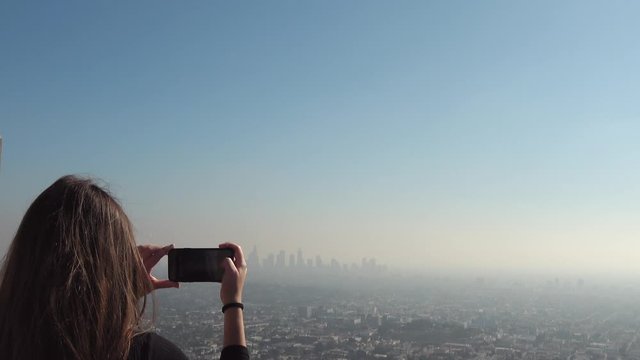4K. Young woman photographs downtown Los Angeles in the distance at the Griffith Park Observatory. The fog makes the buildings look more interesting. Woman makes photos of the Los Angeles skyline. 
