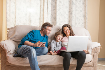 Child, a woman and a man sitting on a sofa watching a laptop and a smart phone.