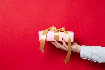 Female hand holds a gift wrap wrapped in pink paper and a gold ribbon bow on a red background