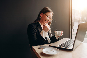 a beautiful girl wearing in office clothes with sunglasses sitting in the cafe with laptop and phone on the table and holding a cup of coffe