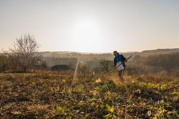 Young handsome man mows dry grass, amazing autumn lanscape with sunset on the background