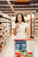 Daily Shopping. Young woman in the supermarket walking with cart reading article on smartphone concentrated