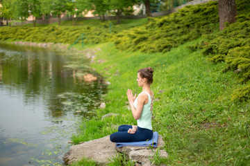 A young sports girl practices yoga on a green lawn by the river, yoga assans posture. Meditation and unity with nature