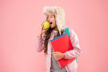 school break. small happy girl earflap hat eat apple. winter holiday and vacation. teen kid pink background. pupil daily life. Modern education. schoolgirl with books after lesson