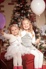 Fototapeta na wymiar two girls with white and purple balloons are sitting on a red ottoman in Christmas decorations