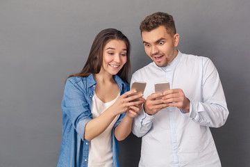 Using Devices. Young couple studio standing isolated on grey showing each other media on smartphone smiling excited