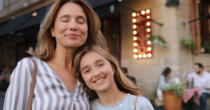 Portrait shot of the cute Caucasian teen girl standing together with her mother at the street and they smiling happily while looking at each other and then turning faces to the camera. Close up.