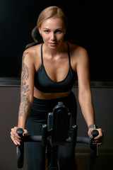 Young confident woman athlete exercising on bicycle indoors. Attractive determined fitness girl doing cycling exercises in dark gym. Functional workout training of sports girl. Cardio training