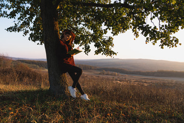 Close up portrait of young brunette girl in red sweater that leaned on a big tree and reading a book, colorful forest on the background