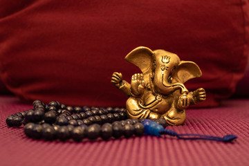 Golden Ganesha statue with blue mala beads on yoga mat in front of a yoga pillow. Close up of Meditation accessories.