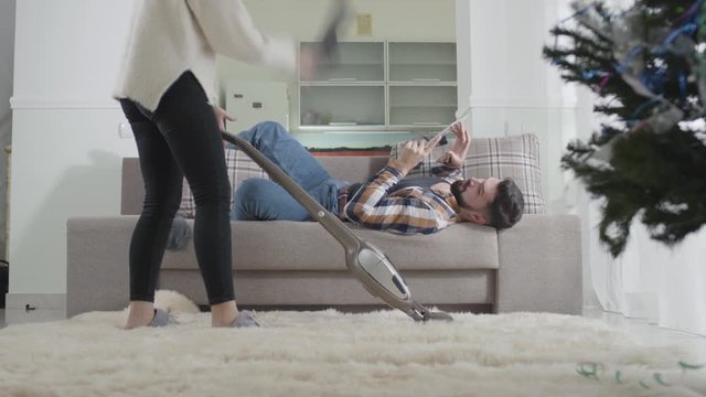 Unrecognizable young Caucasian woman vacuuming carpet at home when her husband lying on the couch. Wife throwing dirty sock on husband and leaving the room. Housework, support, marriage.