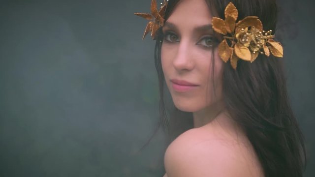 Brunette girl looks back, long hair fly waving in motion. attractive cute face with gentle smile. Backdrop thick fog smoke. Greek goddess with golden laurel wreath white vintage dress. Elven queen