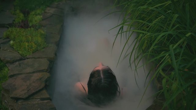 Back view. Sexy woman brunette woman with wet hair lies in stone vintage pool with white steam. Medical wellness treatments. Hot spring spa. Mysterious seductive girl shooting from back without face
