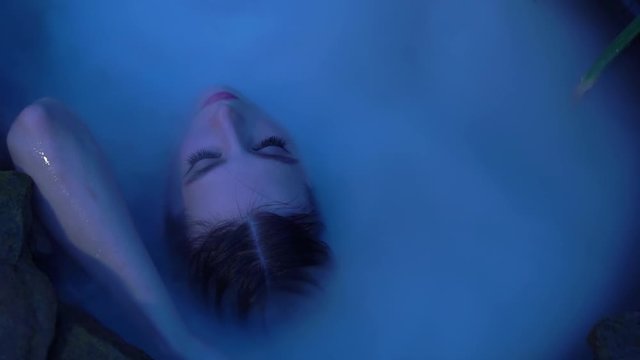 Portrait of gorgeous attractive brunette girl with elegant waterproof makeup. Woman in old stone bath with milk and steam on nature at evening blue dusk. Fashion shooting, sexy girl stroking her face