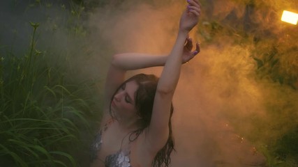 Fashion model brunette girl posing in small pool with hot springs. Slender seductive body. creative silver swimsuit with scales. Backdrop steam fog clouds. Backstage to art of glamorous photoshoot