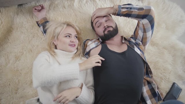 Top view of pretty blond Caucasian woman in white sweater lying with boyfriend or husband on soft carpet and tickling man. Happy playful spouses resting on weekends at home. Leisure, resting.