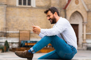 Handsome caucasian smiling bearded hipster sitting outdoors and typing message on smart phone. In background is rustic building.