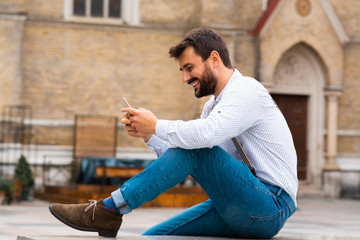 Handsome caucasian smiling bearded hipster sitting outdoors and typing message on smart phone. In background is rustic building.