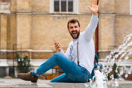 Handsome caucasian bearded fashionable man sitting on funtain, holding smart phone and waving to a friend. In background is rustic building.