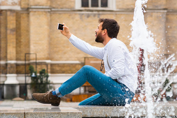 Fashionable caucasian tourist sitting on fountain, sticking tongue out and taking selfie. In background is rustic building.