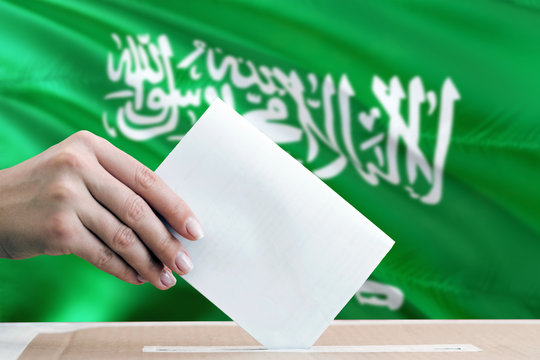 Saudi Arabia Election Concept. Side View Woman Putting A Ballot In A Box On National Flag Background.