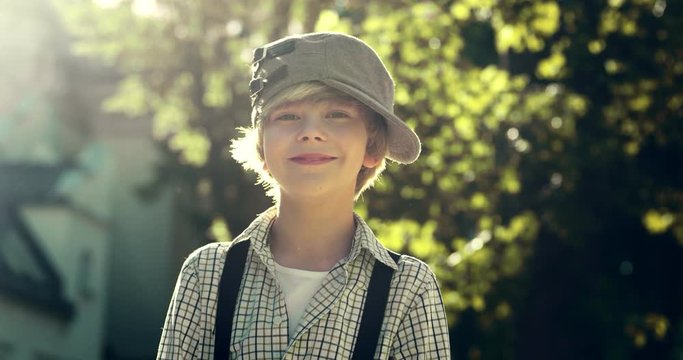 Portrait shot of the good looking happy Caucasian teen boy in hat and suspenders smiling joyfully to the camera outdoors in summer.