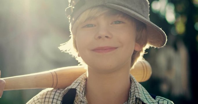 Portrait of the little Caucasian cute teen hooligan boy in a hat posing to the camera and holding a bat over his shoulder. Close up. Outdoor.