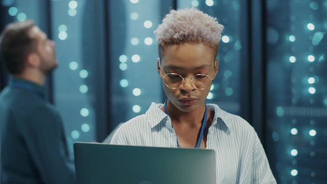 African young woman IT expert with laptop coworking in server corridor. Portrait smart black female engineer standing near secure server racks in data center.