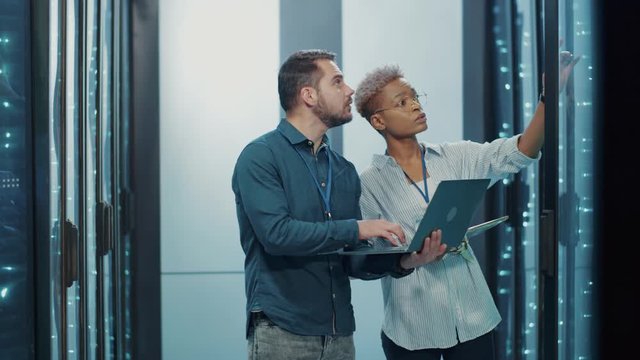 In data center, diverse male and female specialists managing network data server cabinet, running maintenance on laptop, and discussing their work. Professional team.