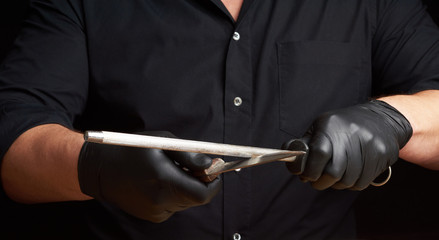 chef in a black shirt and black latex gloves sharpen a kitchen knife on an iron sharpener