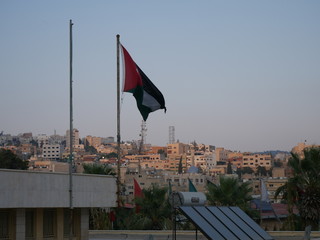 Cityscape of Jerash, Jordan, grey panorama of a modern Arabic city with improvised houses on a hill between few green trees and a tall flag of the country under the blue sky