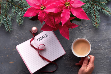 Christmas goals creative flat lay. Hand holding coffee, notebook with natural Xmas decorations, ribbon bookmark and pink trinket. Vibrant pink poinsettia plant and fir twigs on dark background.