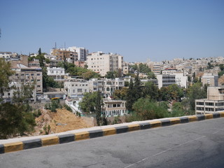 Cityscape of Amman, capitol of Jordan, grey panorama of a modern Arabic city with improvised houses on a hill between few green trees and a tall flag of the country under the blue sky