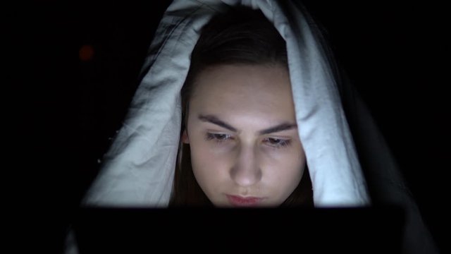 A young woman lies under a blanket with a tablet in her hands. A woman in the dark uses a pad. Modern electronic tablet. Close-up