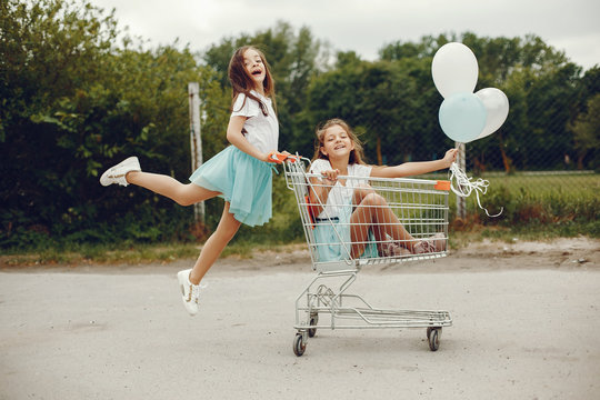 two cute little girls in white T-shirts and blue skirts play in the summer park with balloons and ride in the grocery cart
