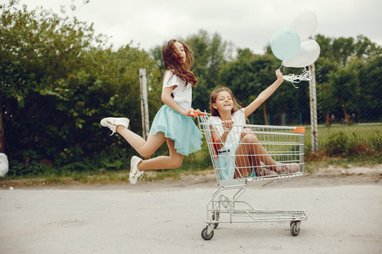 two cute little girls in white T-shirts and blue skirts play in the summer park with balloons and ride in the grocery cart