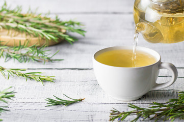 White cup of healthy rosemary tea pouring from teapot with fresh rosemary bunch on white wooden...