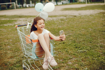 cute little girl in white T-shirt and blue skirt with phone play in the summer park with balloons...