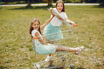 Naklejka na ściany i meble two cute little girls in white T-shirts and blue skirts play in the summer park ride in the grocery cart