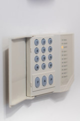 A regular security systems keypad with buttons and a plastic flap mounted on a white wall