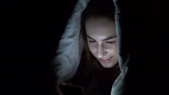 A young woman lies under a blanket with a phone in her hands. A woman in the dark uses a pad. Modern electronic tablet. Close-up