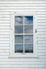 A tall white wooden window with eight panes of glass. The blue sky with clouds is reflecting in the small panes of glass.