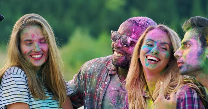 Close up of the joyful young mixed-races male and female friends laughing and celebrating holi holiday outdoors while posing to the smartphone camera and taking selfies photos.