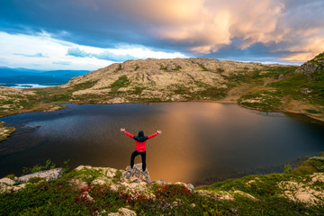 Man with red jacket celebrates with hands raised towards dramatic pink rain clouds and lakes outdoor in the wilderness in norway. Hiking and adventure concept.