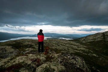 A hiker with red jacket and his dog are standing on a mountain top looking towards lakes and dramatic stormy clouds during blue hour. Hiking and active lifestyle concept.