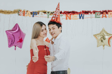 Happy Asian couple celebrating new year together, holding sparklers with New Year backdrop.